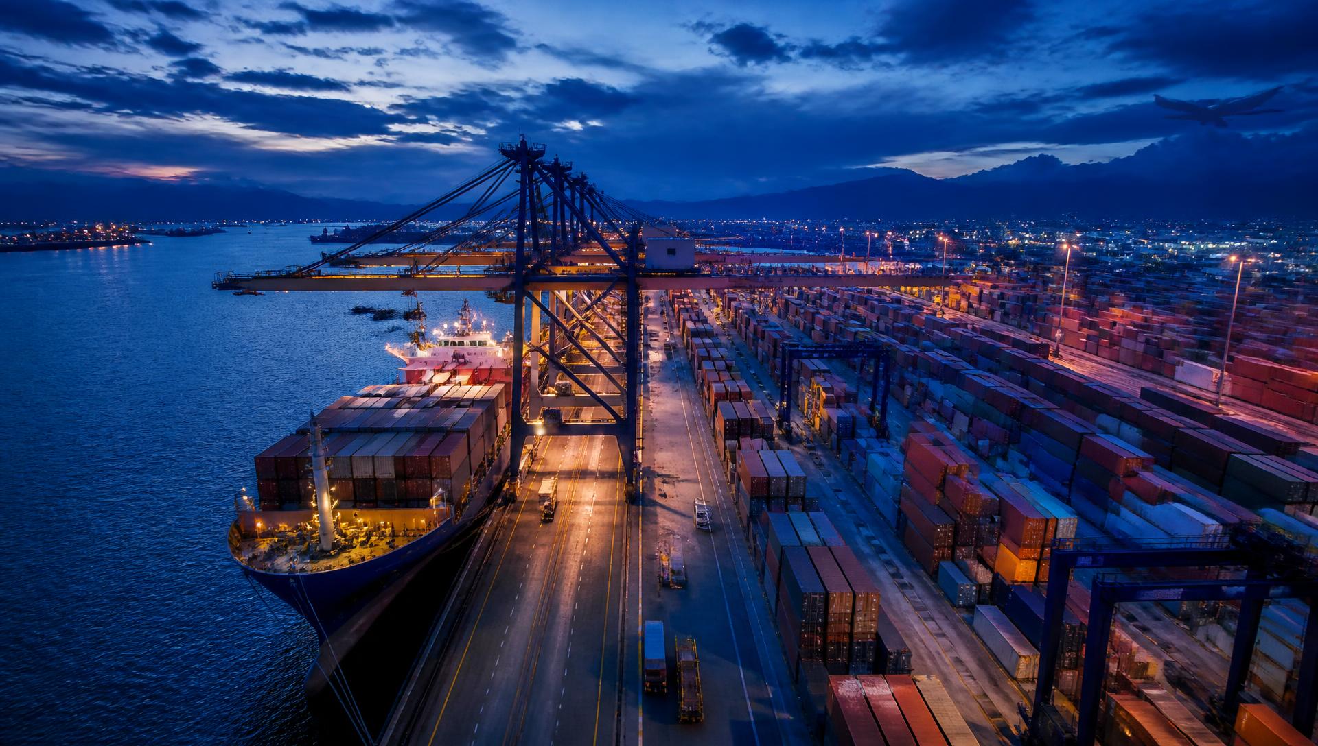 Aerial view of a container port terminal at dusk with a berthed cargo ship, gantry cranes and stacked containers